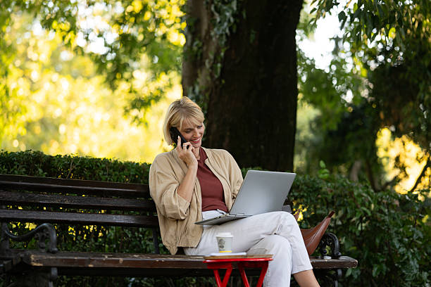 istock Woman sits on a bench using a laptop and talking on a phone in a park during a sunny day 2253932003