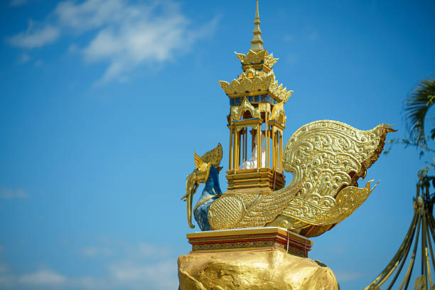 Statue of a Thai mythical creature, Nok Hatsadiling (elephant-headed bird), carrying a golden Busabok pavilion with a small Buddha image on its back. The ornate sculpture features golden wings and a blue body, set against a bright blue sky at a Buddhist temple.