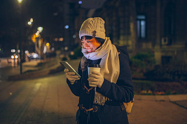 A smiling woman walks through the city at night while holding a smartphone and a takeaway coffee, enjoying an urban evening atmosphere