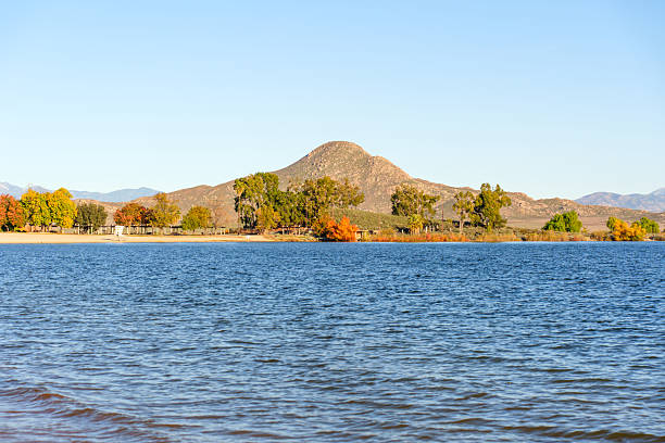 Calm blue waters of Lake Perris reflect the sky, meeting a sandy shore with vibrant autumn trees and a prominent sunlit hill in the background.