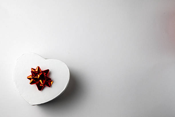 Top view of a white heart-shaped gift box with red ribbon on light background. Minimal romantic composition with copy space, suitable for Valentine’s Day, anniversaries, weddings, celebrations, love concepts, and premium gift packaging.