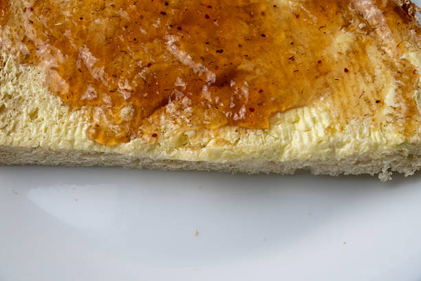Close-up detail of bread spread with margarine and jam, representing simple low-cost food, food insecurity, frugal eating, inflation, and everyday financial hardship. Minimal composition with no people.
