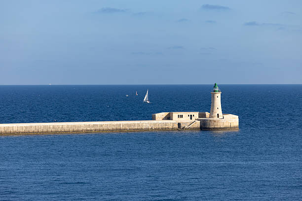 Lighthouse on a long stone breakwater at the entrance of the Grand Harbour in Valletta, Malta. Calm blue Mediterranean Sea with sailing boats in the distance, highlighting maritime navigation and historic coastal architecture.