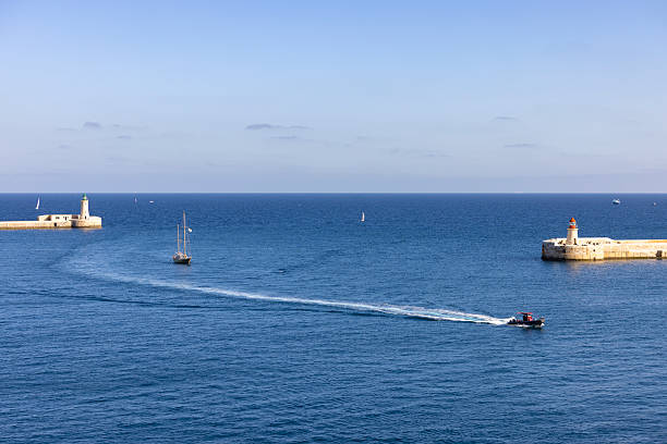 Motorboat creating a white wake while passing between the stone breakwaters at the entrance of the Grand Harbour in Valletta, Malta. Calm blue Mediterranean Sea with sailing boats and historic lighthouses under clear sky.