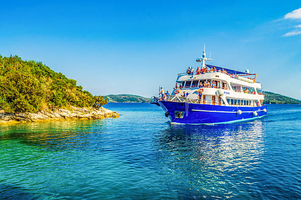 Madouri ,Greece, June 12th 2015:Tour boat navigating by Madouri islet coast and mainland in the distance on bright blue waters and skies on nice summer day.