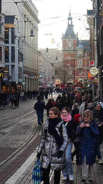 Editorial image taken on December 22, 2025, showing a busy holiday street scene on Leidsestraat in Amsterdam, Netherlands. The cobblestone road with tram tracks is filled with pedestrians dressed in winter clothing, reflecting the pre-Christmas rush. Visible storefronts include Starbucks and La Crêperie, set against a backdrop of mixed architectural styles and a red-brick building with a spire. Overhead tram wires and hanging lamps frame the urban view