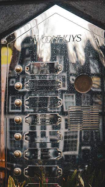 Editorial image taken on December 22, 2025, showing the reflection of traditional Amsterdam canal houses in the metallic surface of an intercom panel shaped like a house. The panel is mounted on a dark brick facade and features nameplates, buttons, and a speaker grille. The mirrored surface captures the distorted outlines of historic buildings, creating a layered visual of urban texture and architectural identity