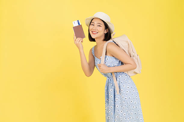 A waist-up studio portrait of an excited young Asian woman ready for an international trip. She is wearing a blue floral summer dress and a straw bucket hat, carrying a beige backpack on one shoulder. She smiles broadly while holding up a brown passport containing a flight boarding pass, isolated against a vibrant yellow background