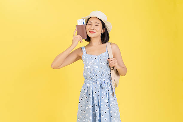 A waist-up studio portrait of an excited young Asian woman ready for an international trip. She is wearing a blue floral summer dress and a straw bucket hat, carrying a beige backpack on one shoulder. She smiles broadly while holding up a brown passport containing a flight boarding pass, isolated against a vibrant yellow background