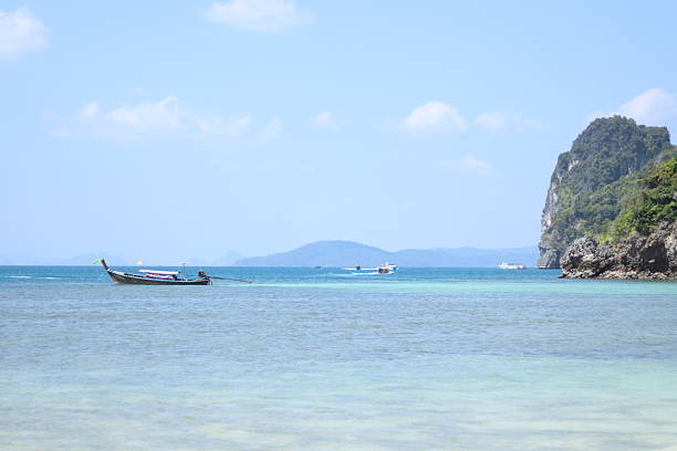 A traditional longtail boat floats on clear turquoise water near a limestone cliff under a blue sky Distant islands are visible across the calm tropical ocean landscape