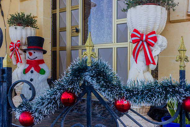 A holiday storefront display with a cheerful snowman, red ribbons, and a garland of evergreen with red baubles draped across a decorative iron gate, complemented by white statue urns.