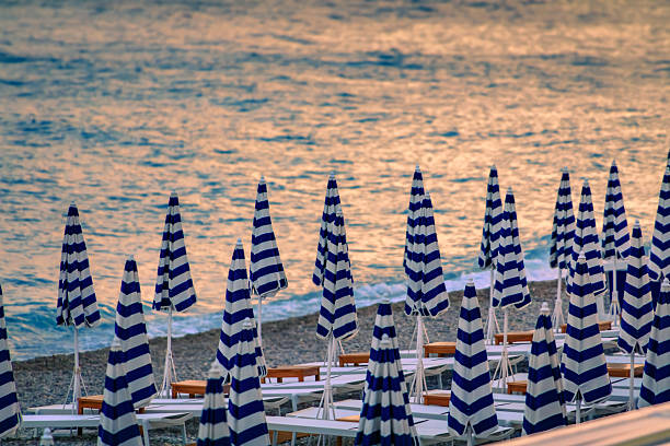 Blue and white striped umbrellas line a pebbled beach during a golden sunset over the Mediterranean Sea along the Promenade des Anglais, Nice, France