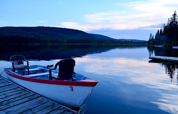 A white and red boat on a glassy calm lake at twilight. Blues and purples relfect in the sky and water, surrounded by forested shoreline.