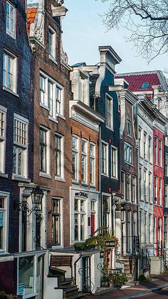 Editorial image taken on December 22, 2025, showing a row of decorative Dutch townhouses on Spiegelgracht in Amsterdam, Netherlands. The buildings feature narrow facades, ornate gables, large windows, and steep staircases. Painted in varied tones - black, white, red, and brown - they reflect the city’s architectural heritage. Wrought iron railings and lantern-style fixtures add historic charm, while bare tree branches hint at the winter season
