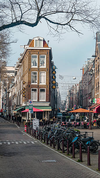 Editorial image taken on December 22, 2025, showing a quiet, empty view of Lijnbaansgracht street in Amsterdam, Netherlands. The scene features traditional Dutch architecture, parked bicycles, and festive street decorations overhead. Outdoor seating from nearby cafés is visible, but no pedestrians are present, emphasizing the stillness of the winter moment