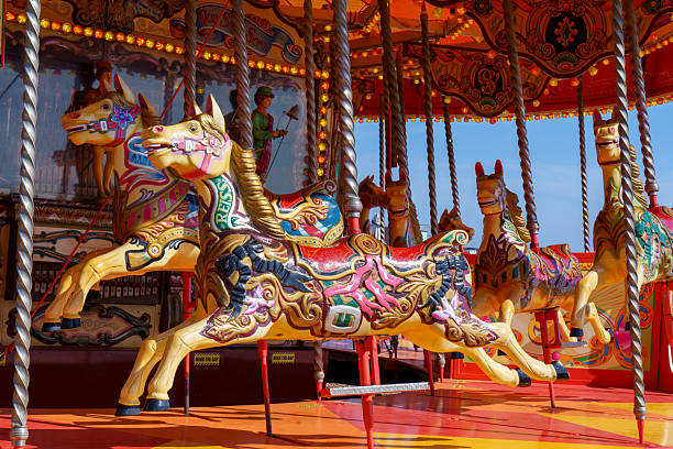A charming seaside amusement spot in Le Treport, Normandy, France, featuring a vintage two-level carousel decorated with ornate panels, pastel colors, and classic fairground horses. Next to it stands a brightly painted snack kiosk with candy-themed illustrations, set against a backdrop of rolling hills, coastal houses, parked cars, and an overcast sky. The grey pebbled ground and muted weather create a nostalgic atmosphere typical of Normandy’s coastal towns. This image suits themes of tourism, leisure, travel, childhood memories, French seaside culture, family attractions, and editorial storytelling, with generous copy space in the cloudy sky and open foreground for versatile layout use without a period