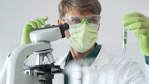 istock Adult male scientist wearing green mask, gloves and goggles using microscope and analyzing sample in test tube in a laboratory, developing new drugs and vaccines. Medicine and science concept 2253972122
