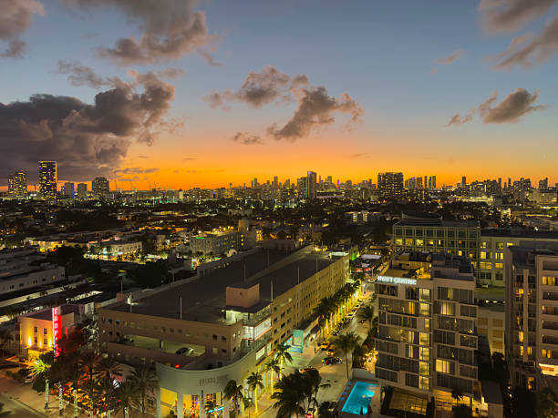 Panoramic view of the Miami skyline at sunset, photographed from Miami Beach, Florida. The city is illuminated by warm evening lights as the sun sets behind the urban horizon, casting golden and orange tones across the sky. Dramatic clouds add depth and atmosphere above the high-rise buildings, while streets, palm trees, and modern architecture fill the foreground.