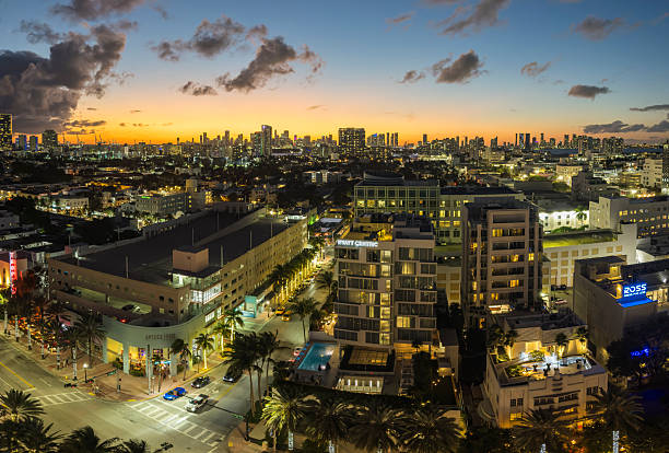 Panoramic view of the Miami skyline at sunset, photographed from Miami Beach, Florida. The city is illuminated by warm evening lights as the sun sets behind the urban horizon, casting golden and orange tones across the sky. Dramatic clouds add depth and atmosphere above the high-rise buildings, while streets, palm trees, and modern architecture fill the foreground.