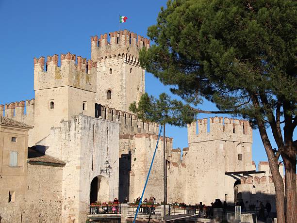 SIRMIONE, ITALY - DECEMBER 29, 2025: Low angle view of the Scaligero Castle, a 14th-century medieval fortress on Garda Lake. The image shows the ancient stone towers and Ghibelline merlons against a clear blue sky, with tourists visiting the historic landmark.
