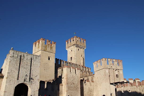 SIRMIONE, ITALY - DECEMBER 29, 2025: Low angle view of the Scaligero Castle, a 14th-century medieval fortress on Garda Lake. The image shows the ancient stone towers and Ghibelline merlons against a clear blue sky, with tourists visiting the historic landmark.