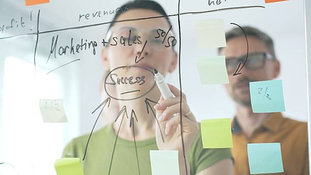 istock Professional team brainstorming, sharing strategy on glass whiteboard with colorful sticky notes, working collaboratively in bright office space. Business people concept 2253980519