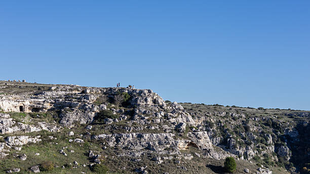 istock panoramic view of ancient rock-cut cave dwellings in a rugged limestone canyon under a clear blue sky, featuring hikers on the cliff edge for scale and perspective. 2253982136