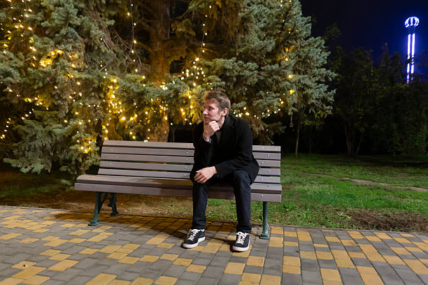 istock A man sits alone on a bench in a park at night surrounded by twinkling lights 2253988902