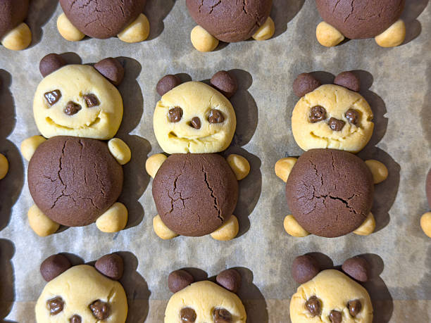 Top view of homemade bear shaped black and white cookies neatly arranged in rows on a parchment lined baking tray. Each cookie features a playful two tone design with chocolate and vanilla dough forming the bear’s face, ears, and body, making this image ideal for themes such as kids desserts, holiday baking, kawaii food, and creative pastries. Vertical composition with repeating pattern and copy space suits cookbooks, blogs, and social media or packaging designs.