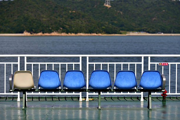 A row of six plastic seats is arranged on a ferry, with five blue seats and one beige seat on the left. The seating is mounted on a metal frame. Behind the seats is a metal railing, and beyond that, a body of water of ocean. In the background, a green, forested hillside is visible across the water. The scene suggests a peaceful voyage with clear weather.