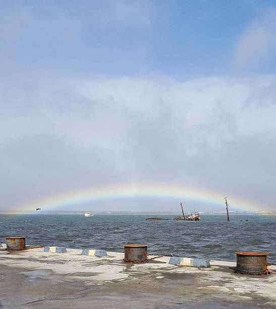 Dramatic post-storm scene in a Kuril Islands bay. A vibrant rainbow arches over a pier, mooring facilities, and a haunting sunken ship. Captures the raw power of nature and maritime resilience, offering a unique blend of beauty and an untold story.
