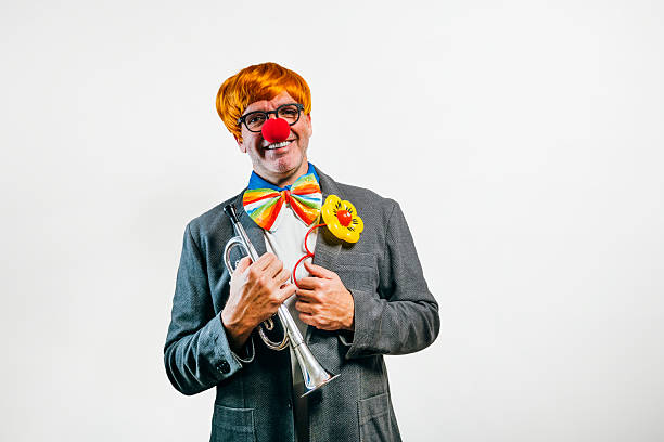 Smiling clown wearing a red nose, orange wig, and colorful bow tie holding a trumpet