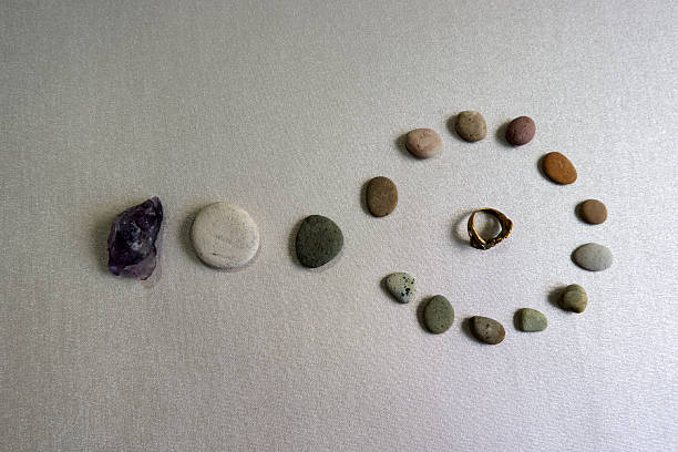 A striking still life composition featuring a rough amethyst crystal, a line of smooth pebbles, and a spiral of smaller stones circling a decorative gold ring. Shot on a reflective silver-grey background, this image conveys themes of spirituality, luxury, natural materials, balance, and the journey of life.
