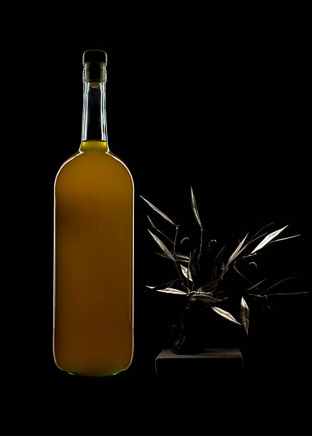 Elegant still life featuring a glass bottle filled with olive oil paired with a decorative olive branch sculpture, isolated on a deep black background. Minimalist composition highlighting Mediterranean aesthetics, premium design, and artistic food presentation.