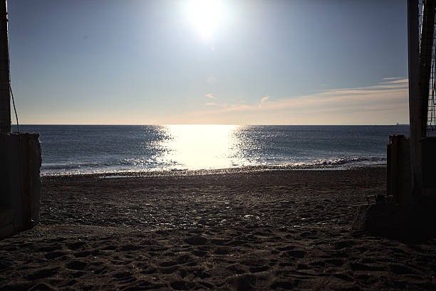 Beach at sunset with sunlight on the water seen from an open gate and through a fence