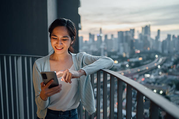 istock A young Asian woman standing on her balcony, using a smartphone with a skyscraper backdrop 2254030479