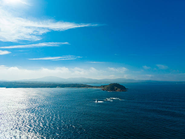 Bright sunlight sparkles on the ocean waves surrounding a peaceful island, with distant mountains softly outlined against the blue sky, creating a picturesque coastal scene.