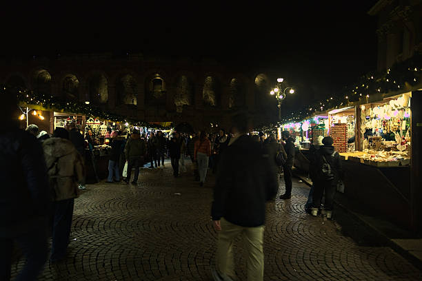 Verona, Italy - 8TH DECEMBER 2025 - Lit Christmas market stalls next to the Arena di Verona and the town hall in Bra square at night, with people passing by, strolling, and browsing stalls