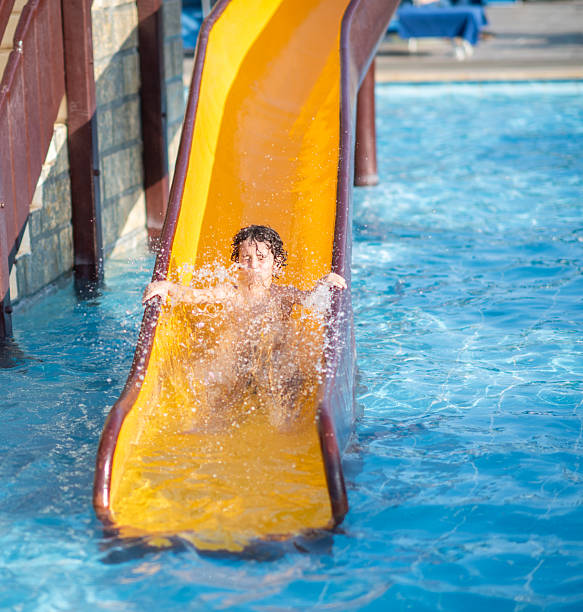 Cute child is enjoying thrilling ride down a water slide, splashing water and having fun in a sunny outdoor swimming pool.