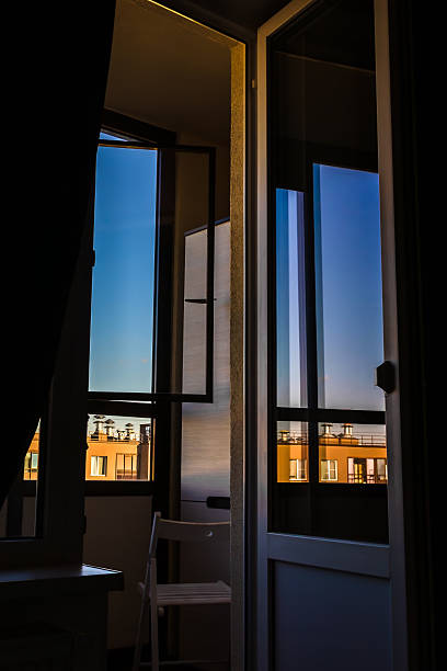 A tall glass door and adjacent window swing open from a dark interior onto a small balcony, framing a warm-lit urban facade under a clear blue dusk sky, with a simple white chair tucked inside.