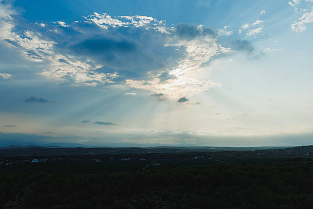 Golden rays of sunlight shine through gray clouds, illuminating the peaceful landscape below. The scene evokes calmness as day transitions to night in the distance.