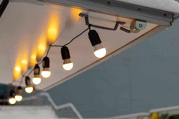 A series of warm, glowing bulbs hang from twisted cables beneath a snow-dusted metal eave, casting a cozy evening light against a softly blurred urban backdrop.