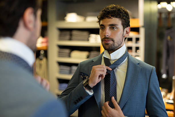 Elegant man adjusting tie in front of mirror while choosing a suit in a luxury menswear store.