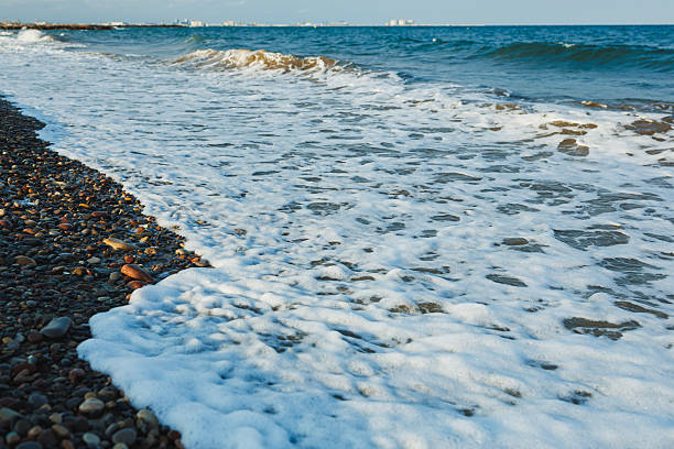 Gentle waves roll onto a rocky beach, forming white foam as they meet the stones under a serene blue sky. The coast stretches far, inviting relaxation and tranquility.