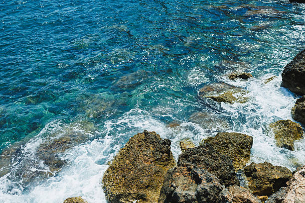 Bright sunlight illuminates the turquoise water as waves crash against rugged rocks at the coast, creating a peaceful scene of nature's beauty, perfect for relaxation and reflection.