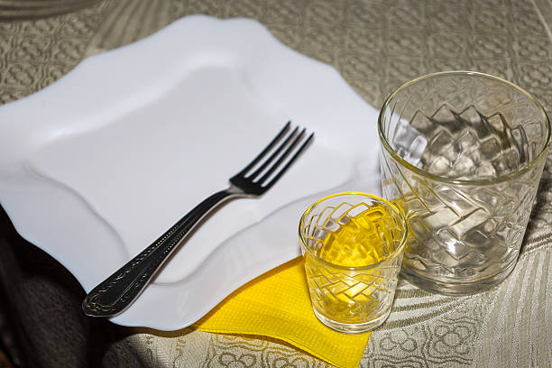A white square plate with a metal fork sits on a table next to a clear glass and a small shot glass on a bright yellow napkin. This minimalist table setting composition against a textured tablecloth creates an atmosphere of preparation for lunch or a fest
