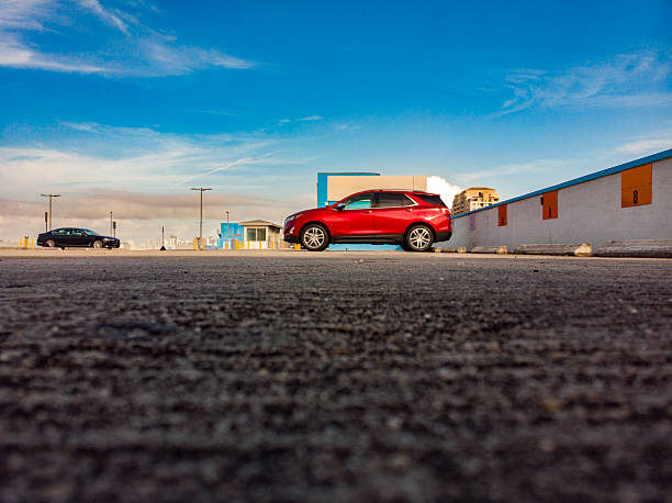 A low-angle perspective captures a red SUV parked on a rooftop parking structure beneath a wide blue sky with soft, wispy clouds. The expansive foreground of textured asphalt emphasizes scale and space, while distant buildings and minimal architectural elements create a clean urban backdrop.