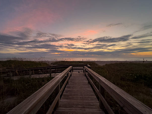 A tranquil sunrise scene at Cocoa Beach, Florida, viewed from a wooden boardwalk leading through coastal dunes toward the Atlantic Ocean. Soft pastel tones of pink, lavender, and gold illuminate layered clouds as early morning light reflects on the calm horizon.