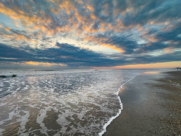 A vibrant sunrise unfolds over the Atlantic Ocean at Cocoa Beach, Florida, as textured clouds glow with soft hues of orange, pink, and blue. Gentle waves wash onto the sandy shoreline, creating reflective patterns in the wet sand that mirror the evolving sky. The wide coastal horizon and dynamic cloud formations convey a sense of calm and natural beauty, capturing the peaceful atmosphere of early morning along Florida’s Space Coast.