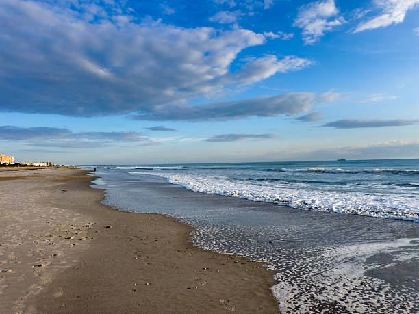 Peaceful early-morning scene along Cocoa Beach, Florida, featuring gentle Atlantic waves rolling onto a wide sandy shoreline under a dramatic blue sky with layered clouds. Soft morning light illuminates the water and wet sand, creating subtle reflections and a calm coastal atmosphere.